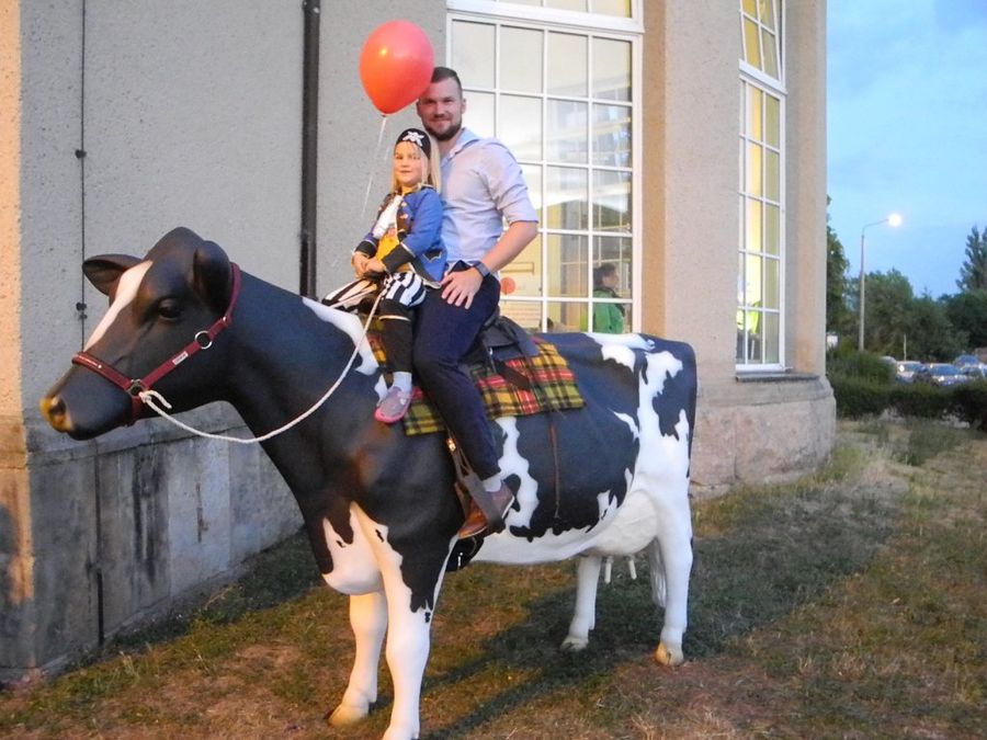 enlarge the image: A life size cow manikin with a saddle in front of the Skills Lab, ridden by a father with his daughter in the moment of the photo