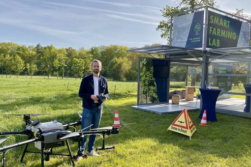 Auf dem Bild ist Wirtschaftsinformatiker Martin Schieck auf einer Wiese neben einer Drohne zu sehen. Neben ihm das Smart Farming Lab.ersuchsfeld. Foto: Diana Smikalla